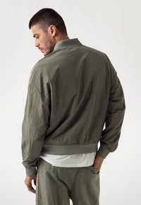 Chaqueta bomber verde oliva en tejido ligero, con cuello y puños acanalados, corte oversize y costuras sin costura.
