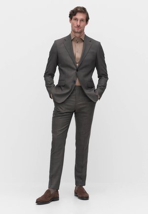 Grijs geruit pak met een getailleerde pasvorm, voorzien van een jasje met twee knopen en een broek met rechte voorkant. Bruin suède schoenen maken de look compleet.