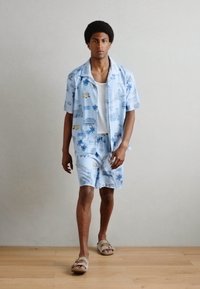Chemise à manches courtes bleu clair et shorts assortis avec un imprimé tropical. La tenue présente une coupe décontractée et est associée à des sandales beiges.