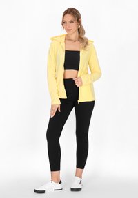 Żółta bluza z kapturem na zamek, zestawiona z czarnym crop topem i czarnymi legginsami. Modelka nosi białe sneakersy z czarnymi akcentami.