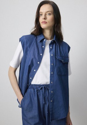 Denim vest with a button-front design, short sleeves, and chest pocket, layered over a white shirt. Soft texture, medium blue color.