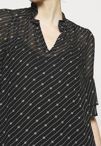 Blusa nera trasparente con scollo a V, colletto arricciato e sottili motivi circolari bianchi lungo strisce bianche diagonali. Maniche corte a campana.