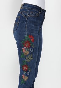 Skinny jeans in dark blue denim feature floral embroidery in red, pink, blue, and green along the left leg, with a standard five-pocket design.