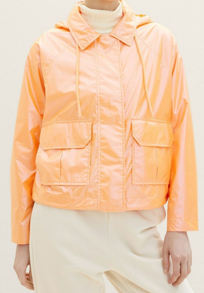 Giacca corta, con cappuccio, lucida di colore arancione chiaro con due tasche frontali. Il materiale sembra liscio e leggero.