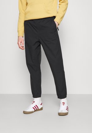 Person iført sorte joggingbukser, hvide sokker, hvide sneakers med røde striber og en gul sweatshirt, stående mod en ensfarvet baggrund.