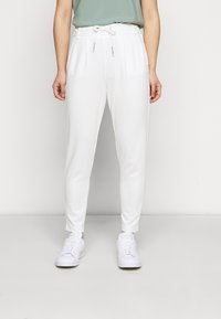 Bílé tapered tepláky s elastickým pasem a šňůrkou, mají boční kapsy a hladkou látku, doplněné o bílé tenisky.