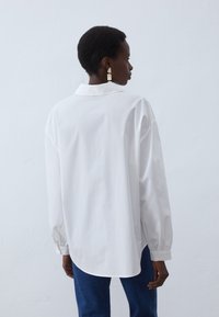 Witte oversized shirt met een puntige kraag, lange mouwen en knoopmansetjes. Vloeiend katoenen materiaal, afgeronde zoom, gedragen met blauwe jeans.