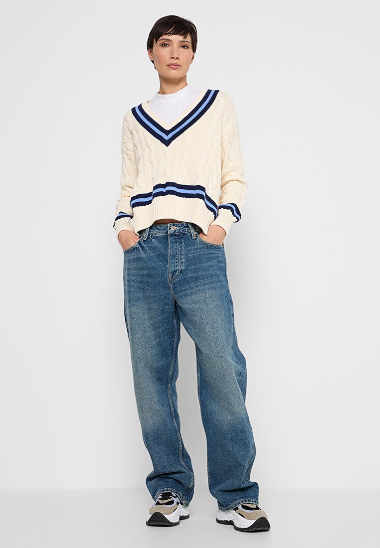 Kremowy sweter w splot warkoczowy z granatowymi paskami przy dekolcie w serek i mankietach, w zestawieniu z luźnymi niebieskimi dżinsami oraz wielokolorowymi trampkami.