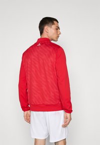 Chaqueta deportiva roja con cremallera y espalda de malla texturizada, puños y cuello acanalados. Pantalones cortos blancos llevados debajo, mostrando un diseño deportivo.