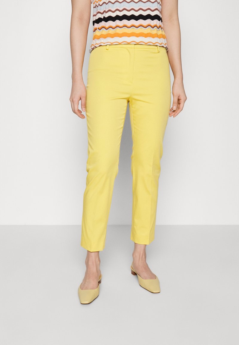 Femme portant un pantalon jaune près du corps, des chaussures beiges à talons bas pointues et un haut sans manches avec des rayures ondulées multicolores.
