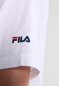 Bílá bavlněná tričko s malým modrým a červeným logem Fila na levém rukávu. Tkanina má hladkou strukturu a klasický kulatý výstřih.