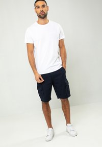 Weißes kurzärmliges Baumwoll-T-Shirt kombiniert mit marineblauen Cargo-Shorts mit Taschen, vor einem schlichten hellen Hintergrund. Weiße Sneakers runden das Outfit ab.