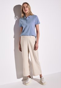Světle modrá polo košile s krátkým rukávem, v kombinaci s krémovými kalhotami širokého střihu a bílými teniskami. Hladká tkanina a minimalistický design.