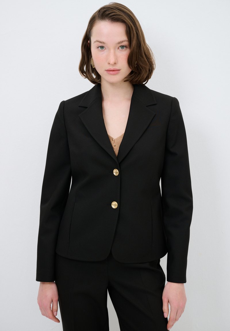 Czarna marynarka o dopasowanym kroju, jednorzędowa, ze złotymi guzikami i wyrazistymi klapami. Wykonana z gładkiego materiału, zestawiona z beżową bluzką.