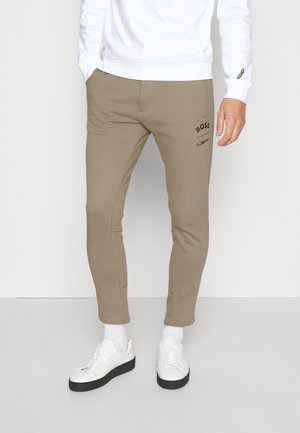 Man draagt beige slim-fit broek met BOSS-logo, witte trui, witte sokken en witte sneakers, staande op een grijze vloer.