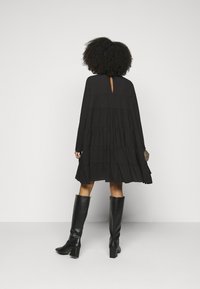 Robe noire à volants avec manches longues et dos découpé en forme de goutte. Le modèle la porte avec des bottes noires montantes jusqu'aux genoux.