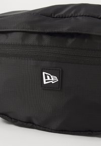 Sac en tissu noir avec fermeture éclair, présentant une surface texturée et un patch logo rectangulaire en blanc et noir.