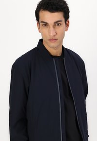 Giacca bomber marina con collo alto, chiusura frontale con zip e accenti grigi a contrasto lungo la zip. Tessuto dalla superficie liscia.