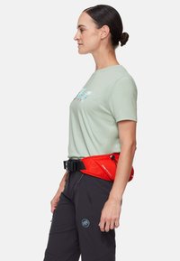 Borsa a vita regolabile rossa con hardware nero, indossata sopra pantaloni grigi. Il modello indossa una maglietta verde chiaro con un'immagine sulla parte anteriore.