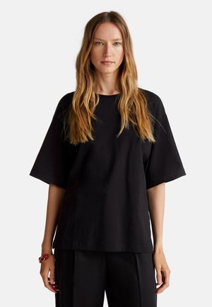 Černé oversized bavlněné tričko má kulatý výstřih a krátké rukávy. Tkanina má hladkou texturu a je kombinována s černými kalhotami.