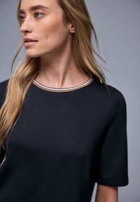 Kobieta z długimi blond włosami, ubrana w czarną bluzkę z krótkim rękawem i okrągłym dekoltem w paski, patrząca w lewo na szarym tle.