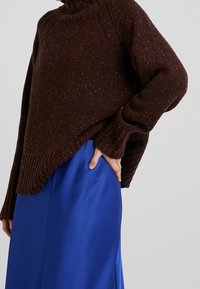 Brunprickig stickad tröja med hög krage, oversized passform, ribbade ärmslut, bärs över en blå slättexturerad kjol, vilket framhäver kontrasten mellan materialen.