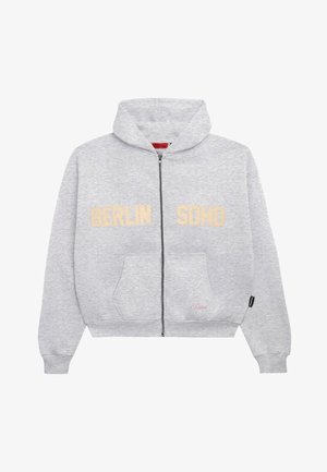 Szürke cipzáros kapucnis pulóver, melyen arany betűkkel "BERLIN SONO" felirat található, kék rövidnadrággal fehér díszítéssel és szürke tornacipővel párosítva. Piros sapka is tartozik hozzá.