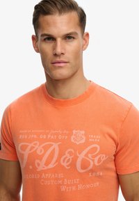 Short-sleeve, bright orange t-shirt made of cotton, featuring white printed text and logo on the front. Smooth texture, classic crew neck design.