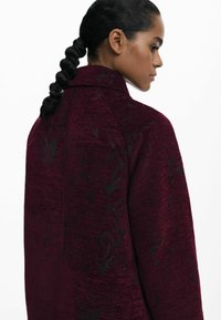 Cappotto bordeaux realizzato in un tessuto testurizzato con un motivo floreale nero, colletto alto e vestibilità strutturata, mostrato da dietro.