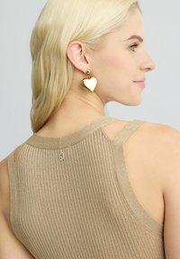 Canotta beige a coste con spalle scoperte e un piccolo dettaglio sul retro, abbinata a orecchini a forma di cuore in oro. Tessuto liscio, design semplice.