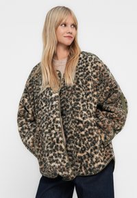 Γυναίκα με ξανθά μαλλιά, ντυμένη με ένα loose, μοτίβο φλις σακάκι με πράσινες λεπτομέρειες, μπεζ πουλόβερ και σκούρα παντελόνια, στέκεται μπροστά σε λευκό φόντο.