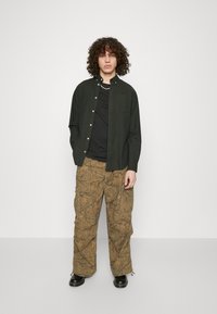 Cămașă neagră cu nasturi peste un tricou negru, asociată cu pantaloni cargo maro texturizați cu un model de frunze și pantofi negri.
