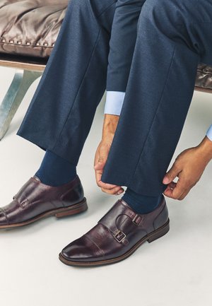 Person iført marineblå habitbukser og mørkeblå sokker, der justerer manchet nær polerede brune double monk strap sko, mens de sidder.