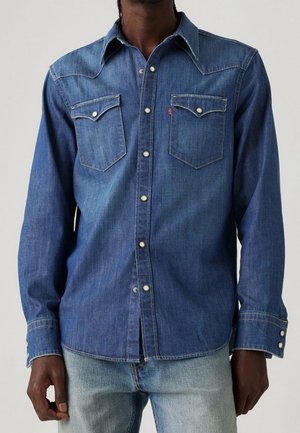 Mann trägt ein geknöpftes, langärmliges blaues Jeanshemd mit zwei Brusttaschen und hellblaue Jeans, steht vor einem einfarbigen Hintergrund.