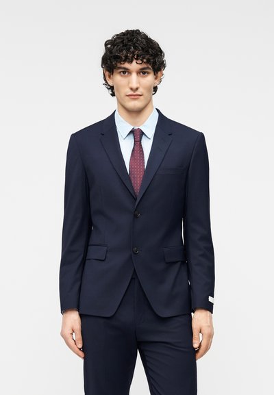 Κοστούμι σε ναυτικό μπλε με δύο κουμπιά, πέτο με σχίσιμο και πλαϊνές τσέπες. Ανοιχτό μπλε πουκάμισο και μπορντό γραβάτα με λευκές πουά. Υφασμα με υφή.