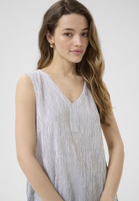 Top sans manches avec un décolleté en V, en tissu blanc avec de fines rayures noires. Texture douce, coupe ample, avec un petit détail de poche avant.