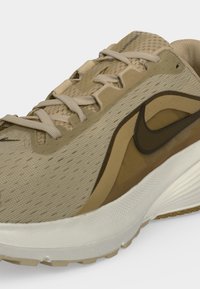 Μπεζ και καφέ αθλητικό παπούτσι τρεξίματος Nike με κορδόνια και λευκή μαλακή σόλα, εμφανιζόμενο από κοντινή πλευρική γωνία.