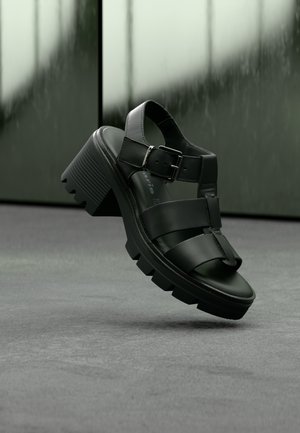 Zwarte leren sandalen met plateauzool en een stevige blokhak, met gekruiste bandjes en een gesp aan de zijkant. Textuur aan de zool voor grip.