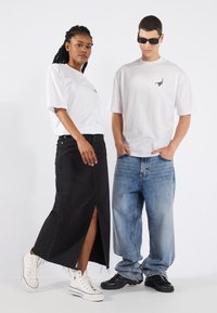 Två modeller bär avslappnade outfits: en vit t-shirt med skorpiontryck, en svart denimkjol med en slit, och lösa blå jeans. Vita och svarta sneakers.
