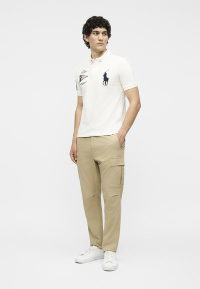 Νέος άνδρας που φοράει λευκό πόλο με λογότυπα και μπεζ παντελόνι cargo, στέκεται με τα χέρια στις τσέπες, κοιτάζοντας προς τα πλάγια.