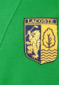 Il tessuto verde a maglia presenta un'etichetta con il logo Lacoste, composta da uno scudo giallo con un leone, un albero e onde, contornato di blu.