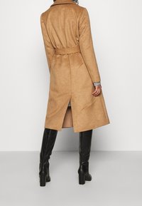 Donna che indossa un cappotto cammello con cintura e spacco sul retro, maniche di un maglione grigio in vista, e stivali neri con tacco al ginocchio, in piedi su un pavimento grigio.