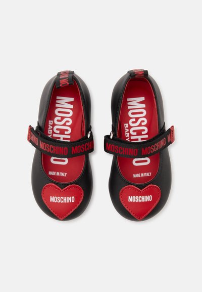 Chaussures bébé noires avec patchs cœur rouges étiquetés Moschino, semelles intérieures rouges et brides noires avec texte Moschino rouge, fabriquées en Italie.