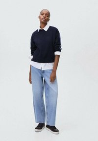 Modra pulover z vojaškim izrezom čez belo srajco s kragnico, v kombinaciji z svetlo modrimi širokimi hlačami in črnimi športnimi copati. Enostaven dizajn z minimalnimi detajli.