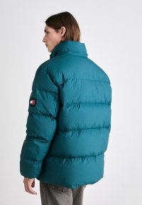 Teal puffer jacket with a high collar, quilted texture, and logo patch on the left sleeve. Features a loose fit and front-facing silhouette.