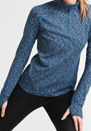 Mujer con una camiseta deportiva de manga larga azul y rosa con estampado y una cremallera corta en el frente, combinada con leggings negros.