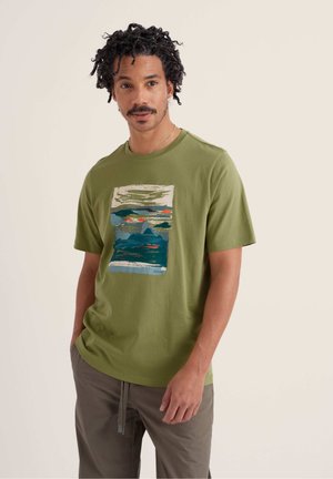 Mees lokkis juustega, kes kannab oliivrohelist T-särki abstraktse maastikugraafikaga ja hallid pükse, seisab beežitaustal.