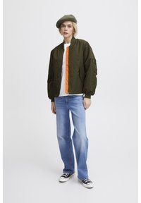 Veste bombardier verte avec fermeture éclair orange, deux poches latérales et une poche sur la manche. Portée sur un t-shirt blanc et un jean bleu clair avec des baskets noires.