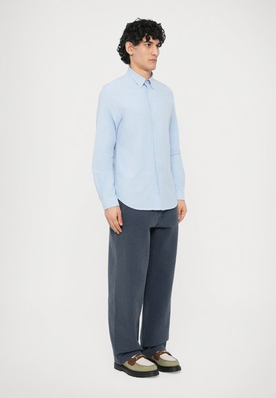 Vaaleansininen nappipaita, pitkähihaiset, yhdistetty tekstuurillisiin, leveälahkeisiin tummansinisiin housuihin ja monivärisiin tennareihin mustilla pohjilla.