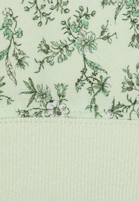 Tessuto floreale verde caratterizzato da fiori delicati e foglie in un verde più scuro. Il materiale ha un aspetto testurizzato e intrecciato, con un bordo inferiore a coste.